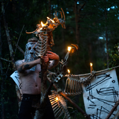 The album cover to Gylne Rune - Uår, A naked man seen from chest up, in a mask with lots of writing on it, holding his hand up. Standing behind a totem made of bones with flames coming up.